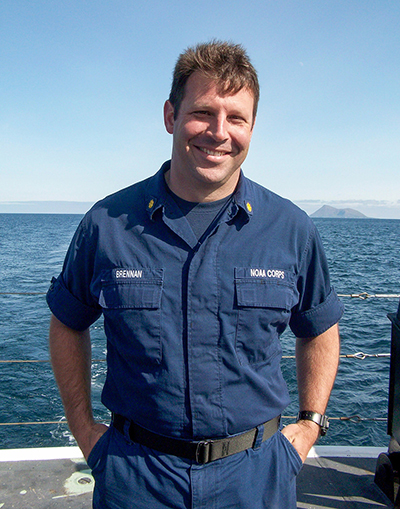 A man with his hands in his pockets stands on the deck of a ship. He is in a blue uniform and is smiling at the camera.