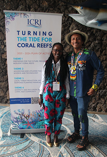 Two people stand next to a sign that reads ‘Turning the tide for coral reefs’. They are dressed in brightly colored clothing and one has a hat on their head.
