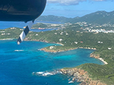 Aerial view of blue water and a green tropical island dotted with buildings, with a plane propeller in frame.