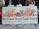 Several people sit in a row behind a table, with a large sign displayed in front of them saying “US Coral Reef Task Force USVI”.