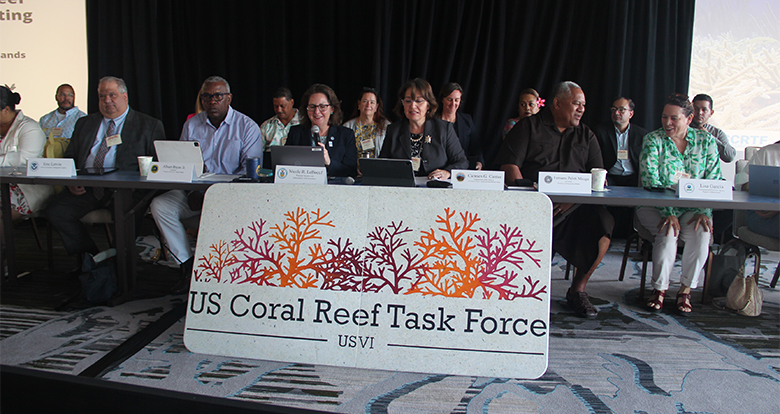 Several people sit in a row behind a table, with a large sign displayed in front of them saying "US Coral Reef Task Force USVI."