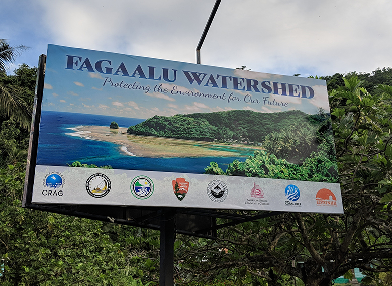A billboard in front of green trees. The billboard has a photo of a watershed and the words Faga'alu Watershed: Protecting the Environment for Our Future at the top and a line of organizational logos along the bottom.