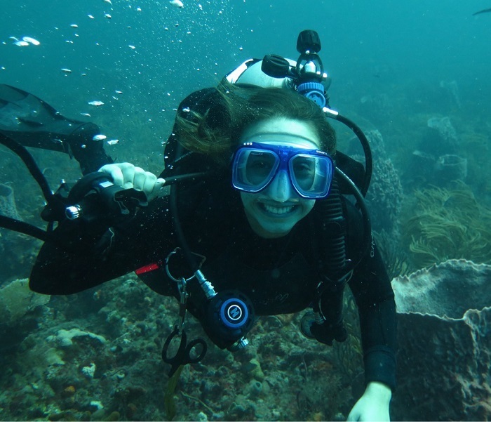 A scuba diver with a blue mask smiles at the camera. They hold the regulator to the side of their face.