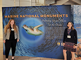 Two women standing on either side of a big poster that reads 'Marine National Monuments' and shows a sea turtle in the center of a school of fish.