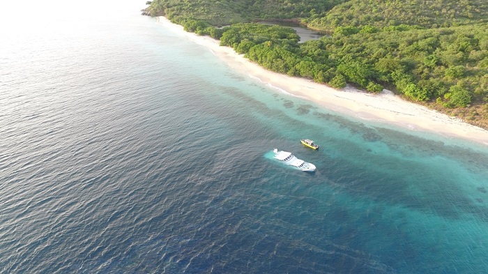 An aerial view of a deserted tropical coastline. A large white boat is halfway submerged in shallow water. A smaller yellow boat sits on the surface next to it.